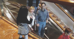 A little girl stands beside an escalator at a shopping mall, waving and saying bye to people as they ride past while adults smile and wave back.