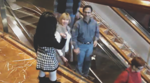 A little girl stands beside an escalator at a shopping mall, waving and saying bye to people as they ride past while adults smile and wave back.