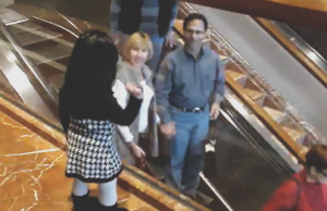 A little girl stands beside an escalator at a shopping mall, waving and saying bye to people as they ride past while adults smile and wave back.