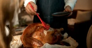 A person wearing an apron brushing glaze onto a roasted turkey indoors, preparing for a Thanksgiving feast