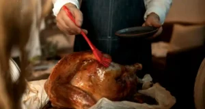 A person wearing an apron brushing glaze onto a roasted turkey indoors, preparing for a Thanksgiving feast