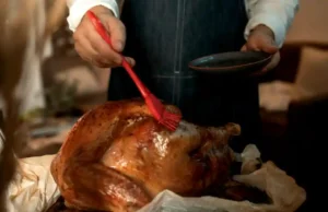 A person wearing an apron brushing glaze onto a roasted turkey indoors, preparing for a Thanksgiving feast
