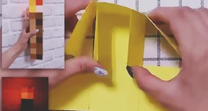 A handmade Minecraft torch made from cardboard and colored paper squares, with a pixelated brown handle and orange-yellow flame top.