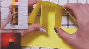 A handmade Minecraft torch made from cardboard and colored paper squares, with a pixelated brown handle and orange-yellow flame top.