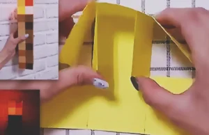 A handmade Minecraft torch made from cardboard and colored paper squares, with a pixelated brown handle and orange-yellow flame top.