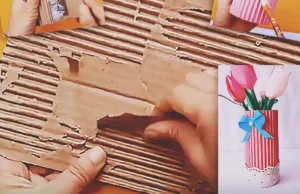 A handmade pink flower vase made from a recycled plastic bottle wrapped in corrugated cardboard, decorated with white lace, a blue bow, and paper flowers.
