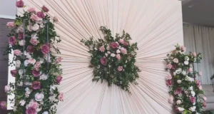 An elegant DIY wedding backdrop made from ivory curtains gathered into a pleated sunburst design with pink rose floral arrangements in the center and on both sides.
