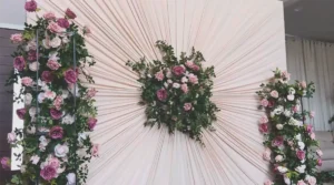 An elegant DIY wedding backdrop made from ivory curtains gathered into a pleated sunburst design with pink rose floral arrangements in the center and on both sides.