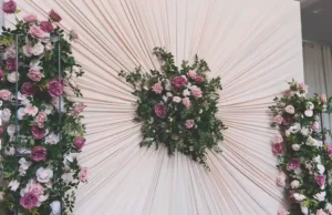 An elegant DIY wedding backdrop made from ivory curtains gathered into a pleated sunburst design with pink rose floral arrangements in the center and on both sides.