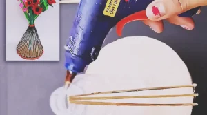 A handmade flower vase made from a recycled plastic bottle, cardboard base, and wooden sticks, painted dark and decorated with red trim while holding colorful handmade flowers.