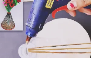 A handmade flower vase made from a recycled plastic bottle, cardboard base, and wooden sticks, painted dark and decorated with red trim while holding colorful handmade flowers.