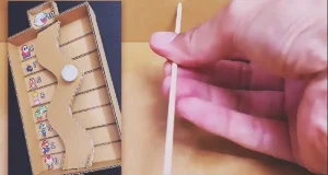 A handmade cardboard balance game with a curved tilting platform, numbered scoring lanes, and a decorated ping pong ball controlled by an external handle made from cardboard and bamboo skewers.