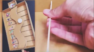 A handmade cardboard balance game with a curved tilting platform, numbered scoring lanes, and a decorated ping pong ball controlled by an external handle made from cardboard and bamboo skewers.