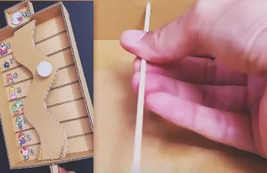 A handmade cardboard balance game with a curved tilting platform, numbered scoring lanes, and a decorated ping pong ball controlled by an external handle made from cardboard and bamboo skewers.