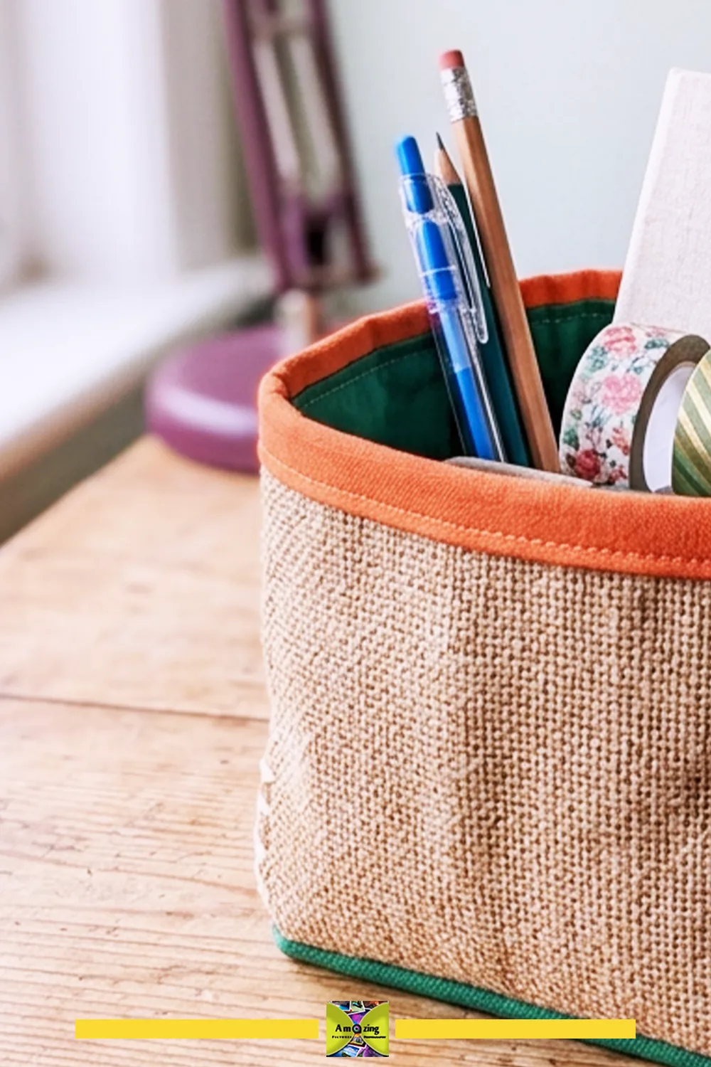 A fabric desktop basket holding pens, a pencil, washi tape, and a notepad on a wooden desk.