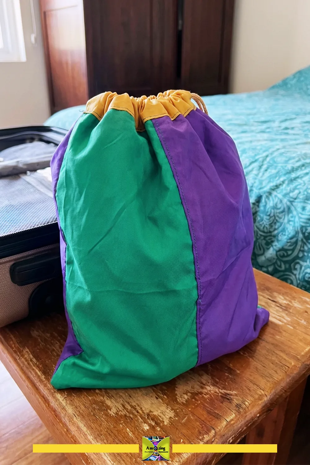 Green, purple, and yellow drawstring shoe-storage bag closed on a wooden table near a suitcase and bed.