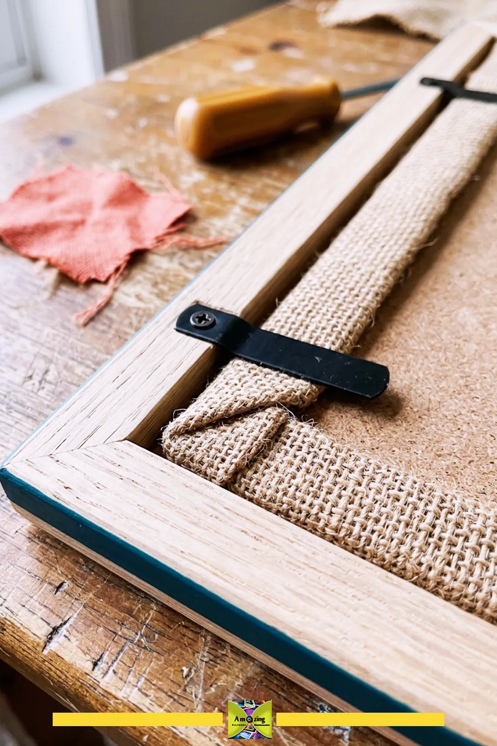 Close-up of a burlap pinboard edge wrapped and fastened inside a wooden frame with a metal clip.