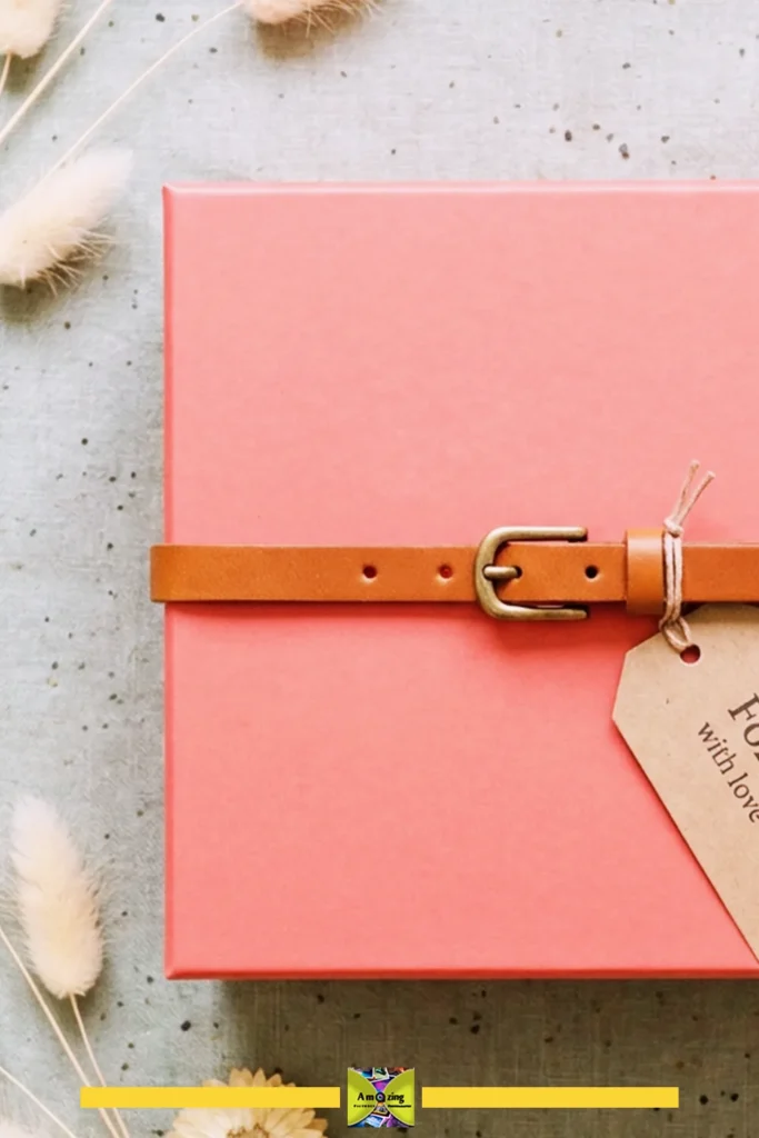 Pink gift box secured with a tan leather band and kraft paper tag on a fabric background