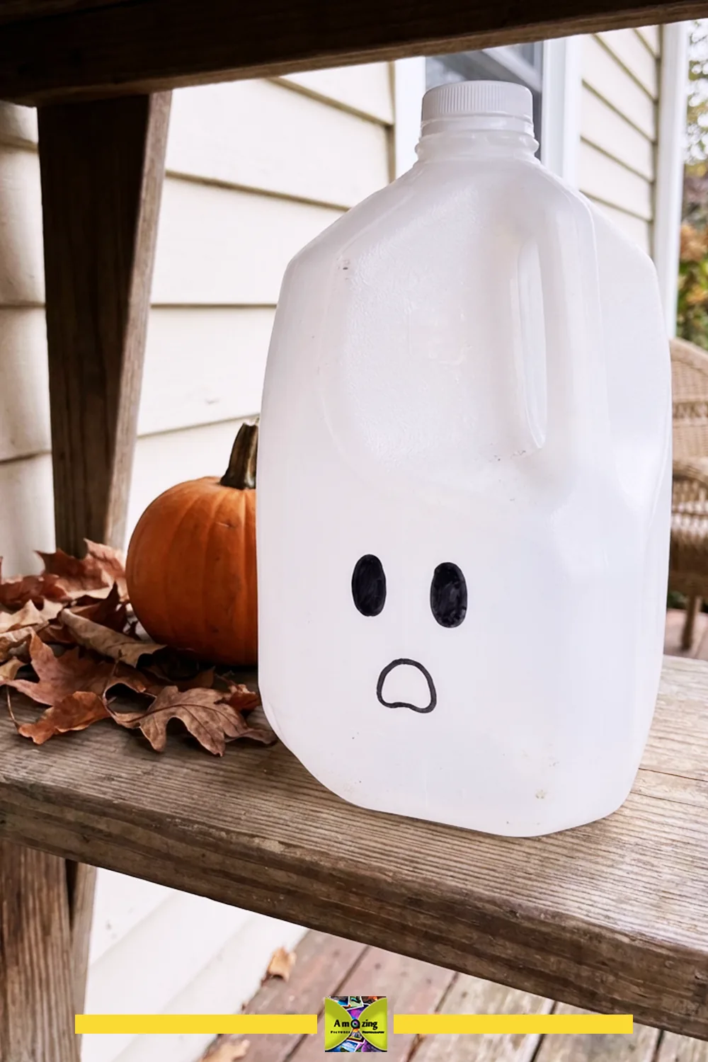 Plastic milk jug decorated as a ghost next to a pumpkin and autumn leaves on a wooden porch shelf.