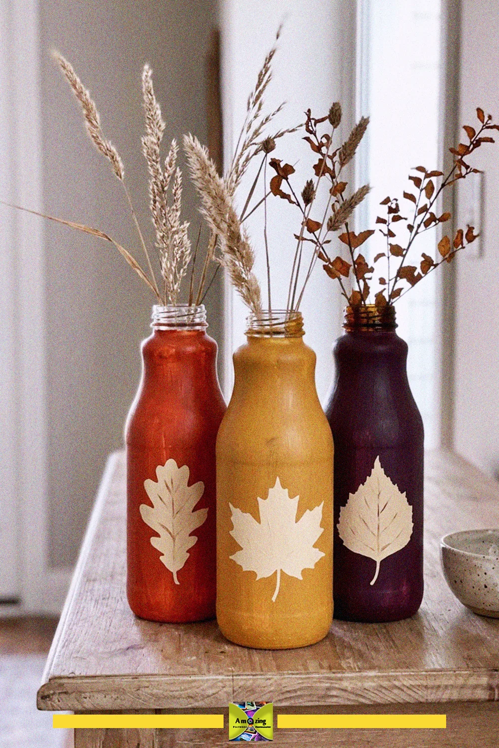 Three upcycled glass bottles painted in rust, mustard and plum with leaf motifs, holding dried grasses for fall decor