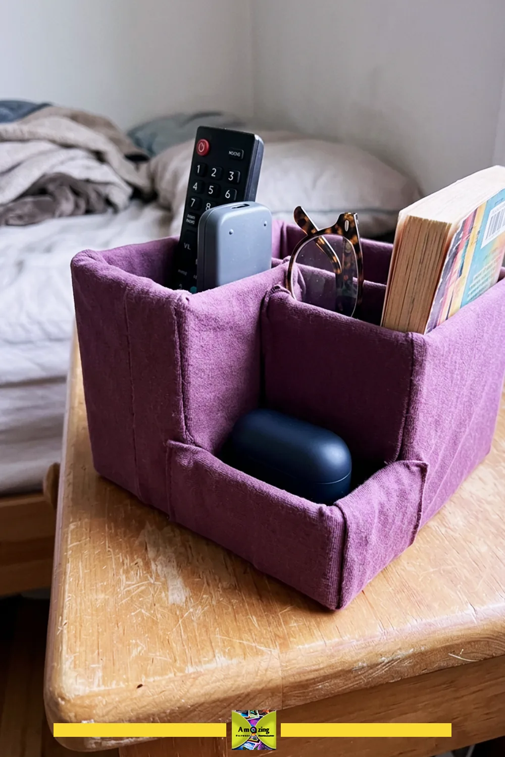 Purple fabric-covered cardboard bedside organizer holding remotes, glasses and a book on a nightstand