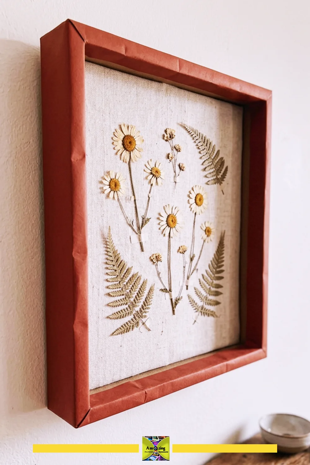 DIY cardboard shadow box frame with pressed daisies and ferns on a fabric background, hanging on a wall