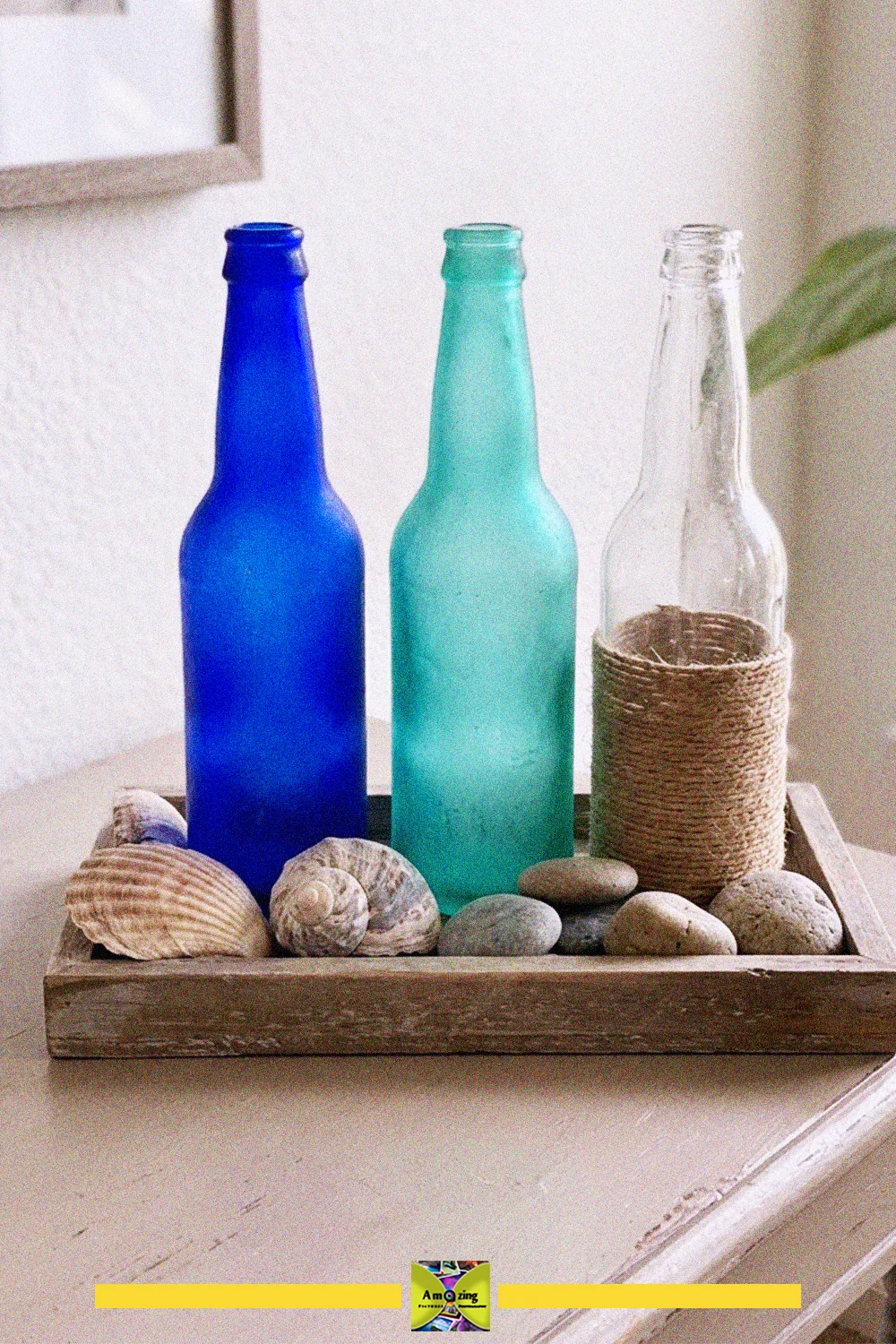 Blue, green and twine-wrapped clear glass bottles with seashells and stones in a wooden tray as coastal decor