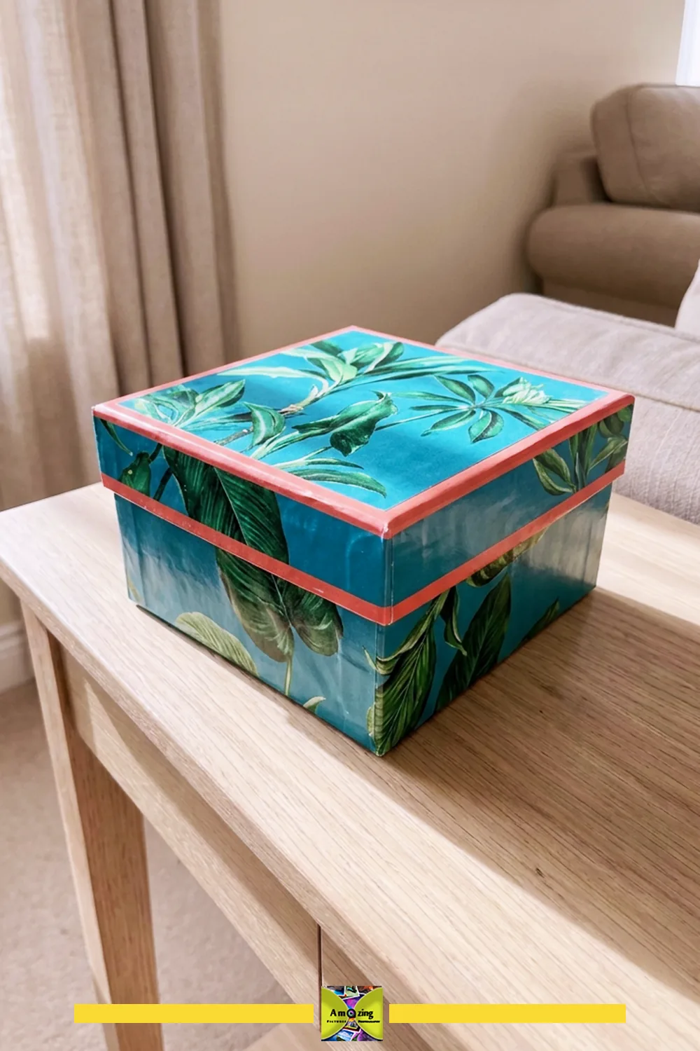Handmade decorative gift box with tropical leaf design on a wooden table in a cozy living room.