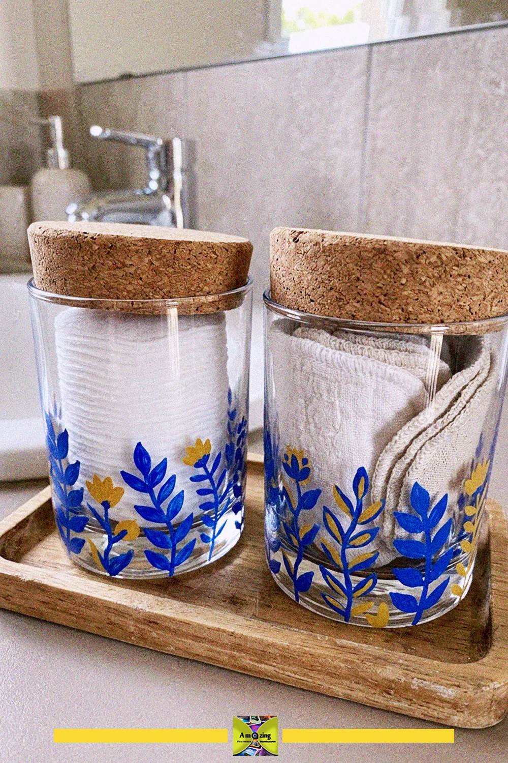 Two upcycled glass cups painted with blue and yellow leaves, with cork lids, holding cotton pads