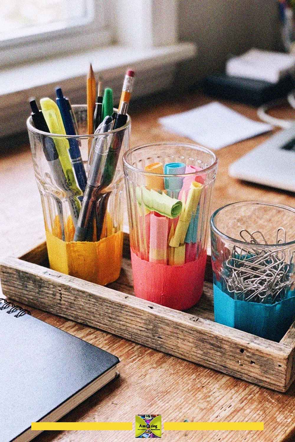 Three mismatched glass cups with painted bases holding pens, pencils, paper clips and notes on a wooden tray