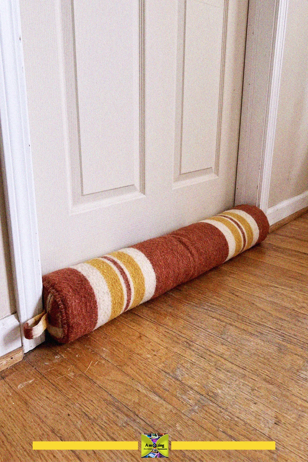 Striped draft stopper made from a recycled blanket lying at the bottom of a white door on a wooden floor