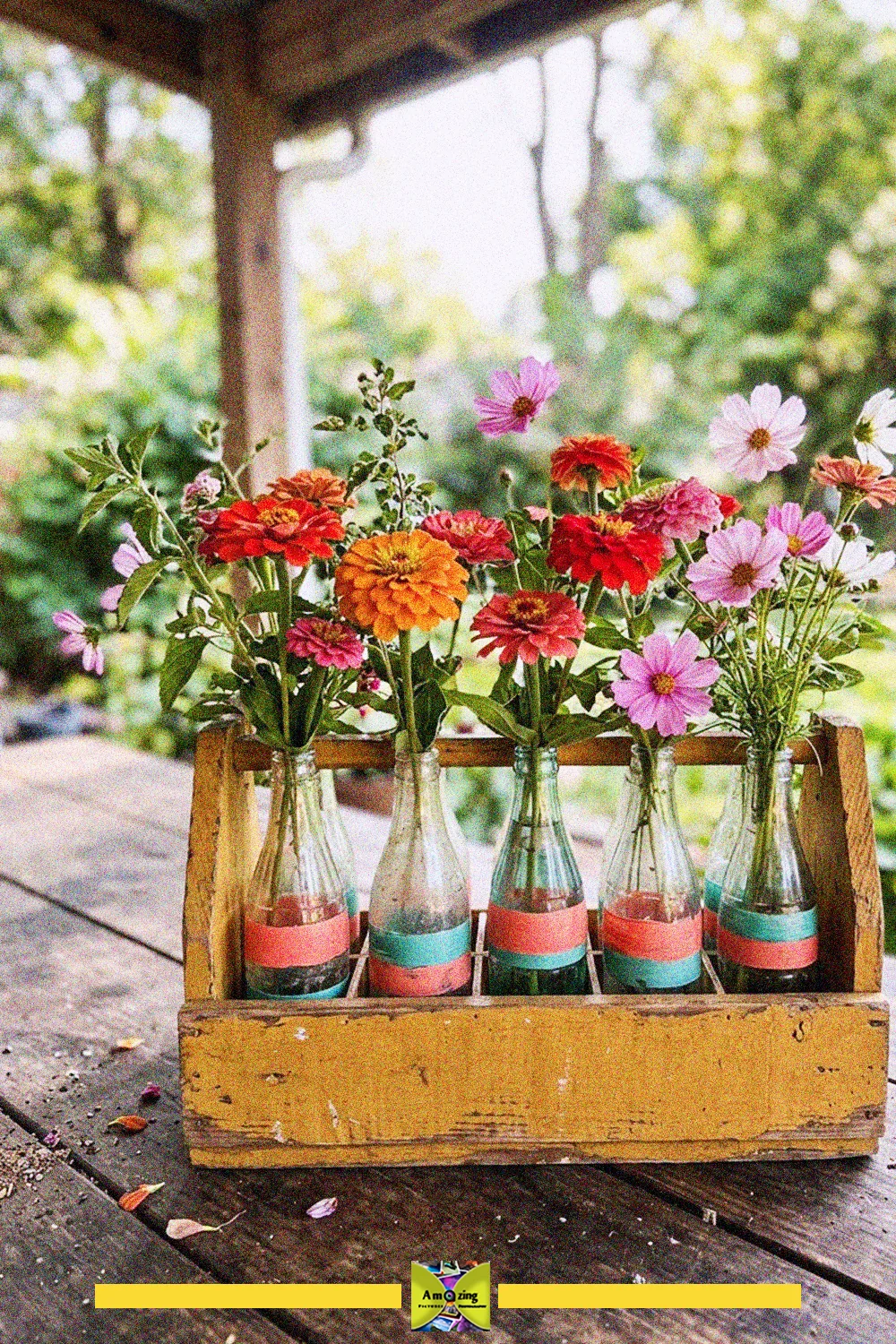 Row of small glass bottles with colorful wildflowers inside a painted yellow wooden crate on a patio table