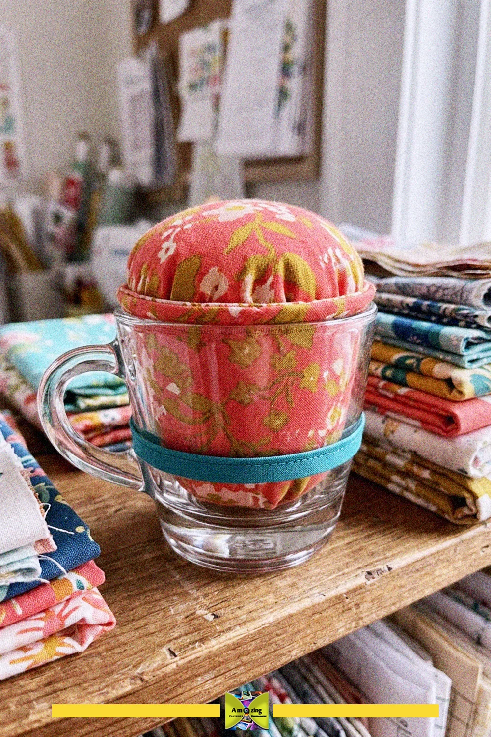 Glass cup pincushion with a domed floral fabric top and teal trim on a shelf beside stacks of fabric