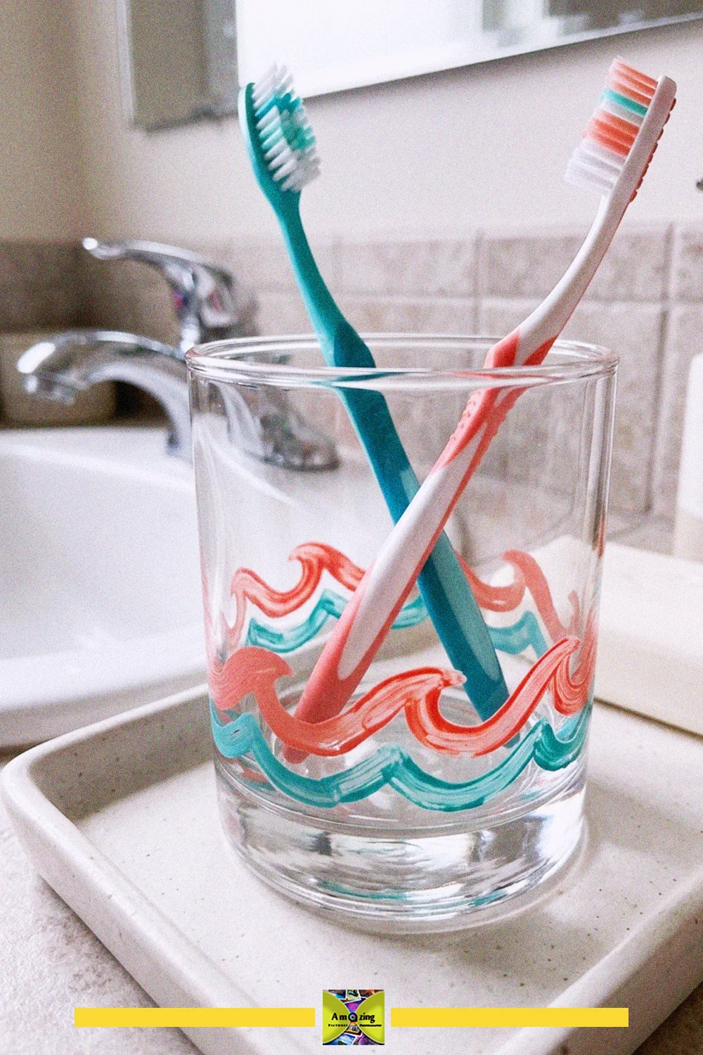 Repurposed glass tumbler painted with coral and teal waves used as a toothbrush holder on a bathroom sink