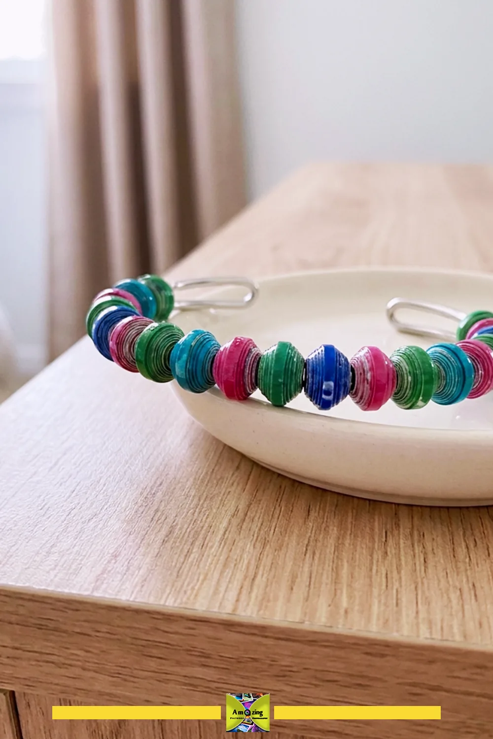 Colorful handmade paper-bead cuff bracelet resting on a ceramic dish on a wooden table.